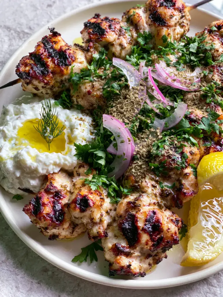 Greek chicken skewers with Authentic Creamy Tzatziki Sauce 2 A macro photo of grilled Greek chicken skewers on an off-white plate with a drizzle of olive oil on creamy tzatziki. (Greek chicken skewers)