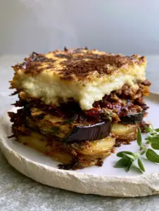 Close up side view of Eggplant Moussaka showing the 2-inch thick bechamel layer and dark red lamb ragu with fresh oregano garnish.