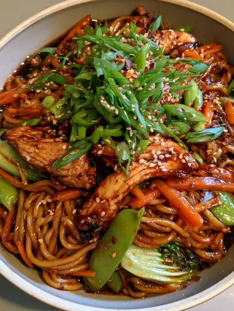 Chicken Lo Mein bowl garnished with green scallions, sesame seeds, and red chili flakes under soft natural light.