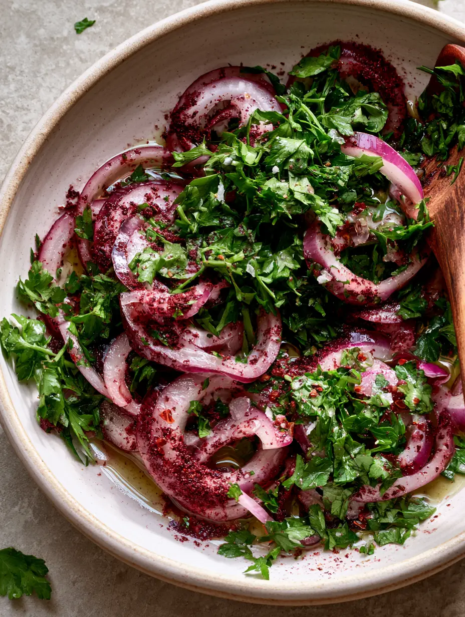Close up of paper-thin red onions massaged with deep burgundy sumac and fresh parsley in a white ceramic bowl. (sumac onion salad)