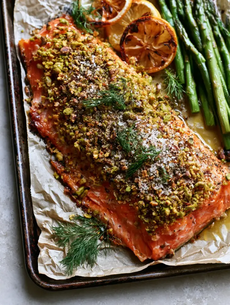 A vibrant rainbow trout fillet next to charred lemon wheels and roasted asparagus on wrinkled parchment. (pistachio-crusted rainbow trout)