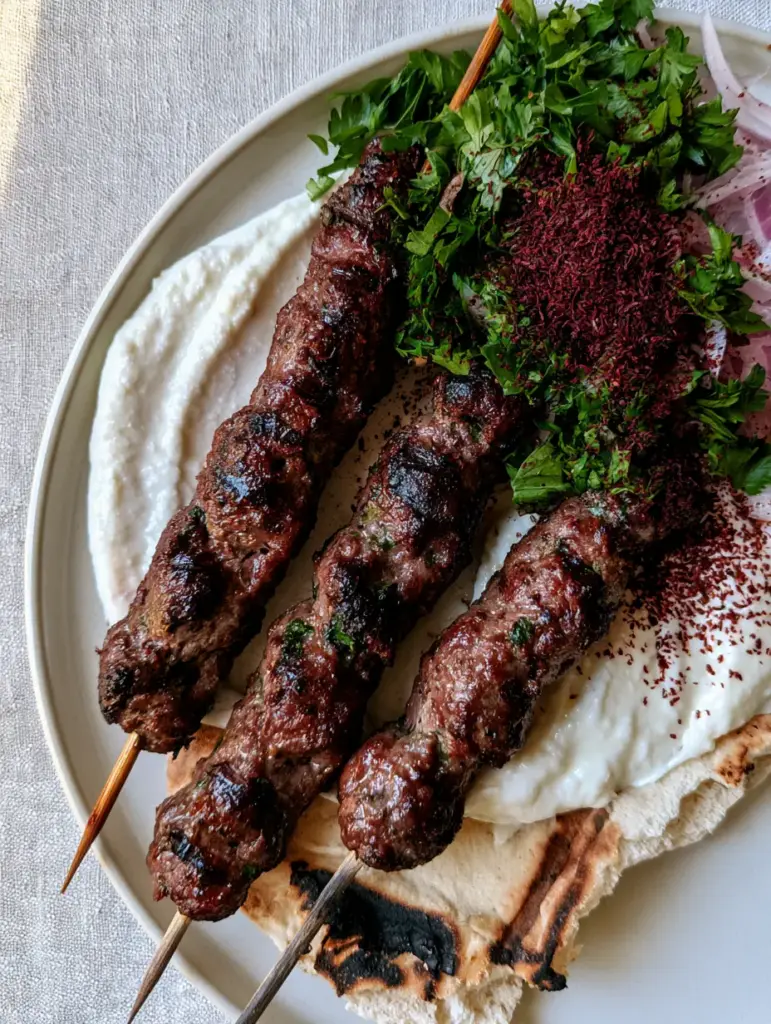 Smartphone photo of Lebanese Kofta skewers with a thick smear of snowy white garlic toum and red sumac dusting.